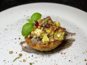 A toasted tartlet topped with golden couscous, diced vegetables, feta, and fresh basil sits on a white plate, sprinkled with herbs and crumbs.