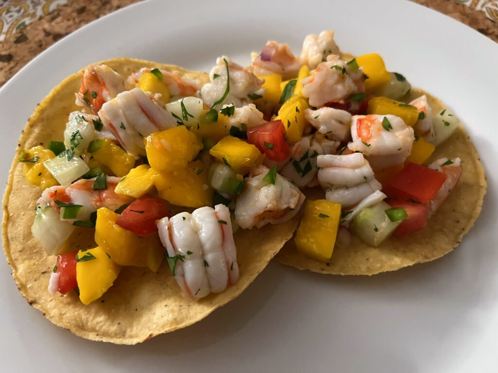 Two crisp tostadas on a white plate are topped with shrimp ceviche, diced mango, tomato, cucumber, onion, and herbs—a colorful, fresh-looking meal.