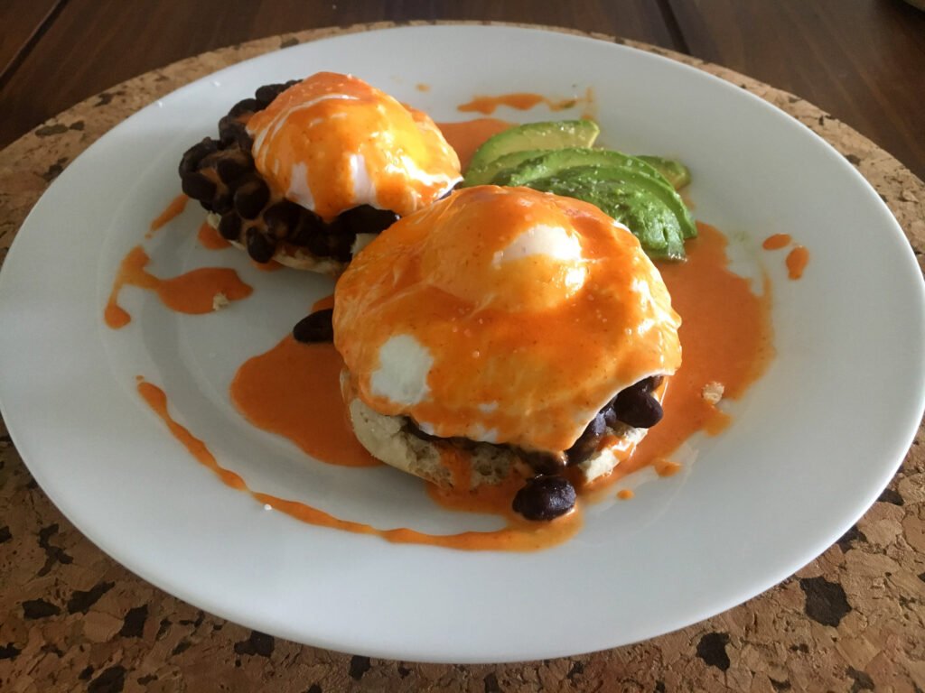 Two English muffin halves topped with black beans, poached eggs, and orange sauce on a white plate, with sliced avocado; a hearty, casual meal.