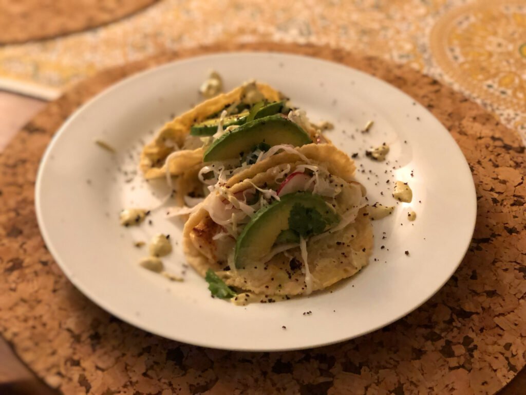 Three soft tacos filled with shredded meat and vegetables, topped with avocado slices and herbs, sit on a white plate; a warm, rustic meal.