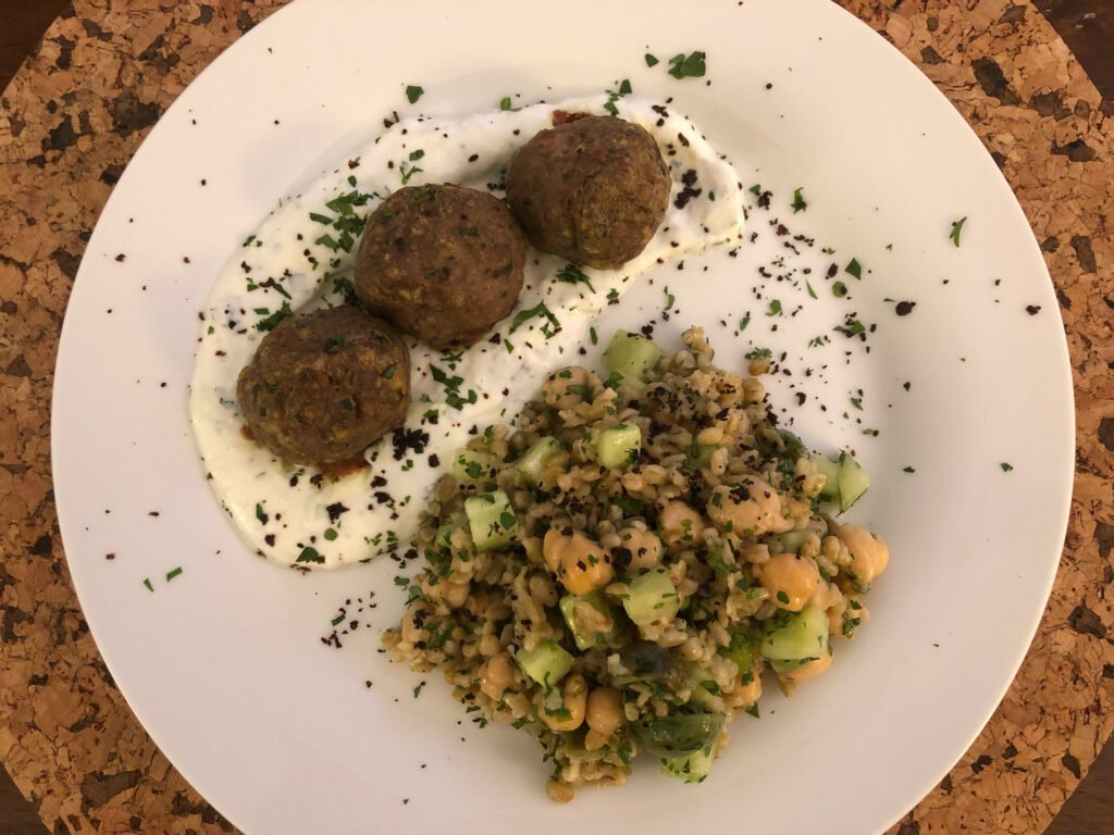 A white plate holds three browned falafel balls on herb-flecked yogurt beside a hearty grain salad with chickpeas and cucumber, presented on a cork table.