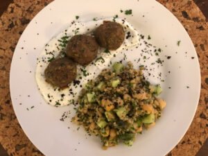 A white plate holds three browned falafel balls on herb-flecked yogurt beside a hearty grain salad with chickpeas and cucumber, presented on a cork table.