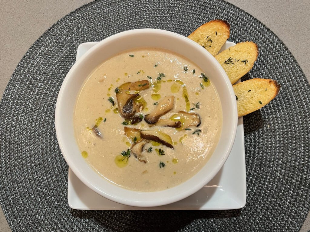 A white bowl of creamy mushroom soup, drizzled with olive oil and herbs, sits on a square plate with three toasted bread slices; warm, inviting presentation.