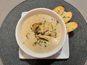 A white bowl of creamy mushroom soup, drizzled with olive oil and herbs, sits on a square plate with three toasted bread slices; warm, inviting presentation.