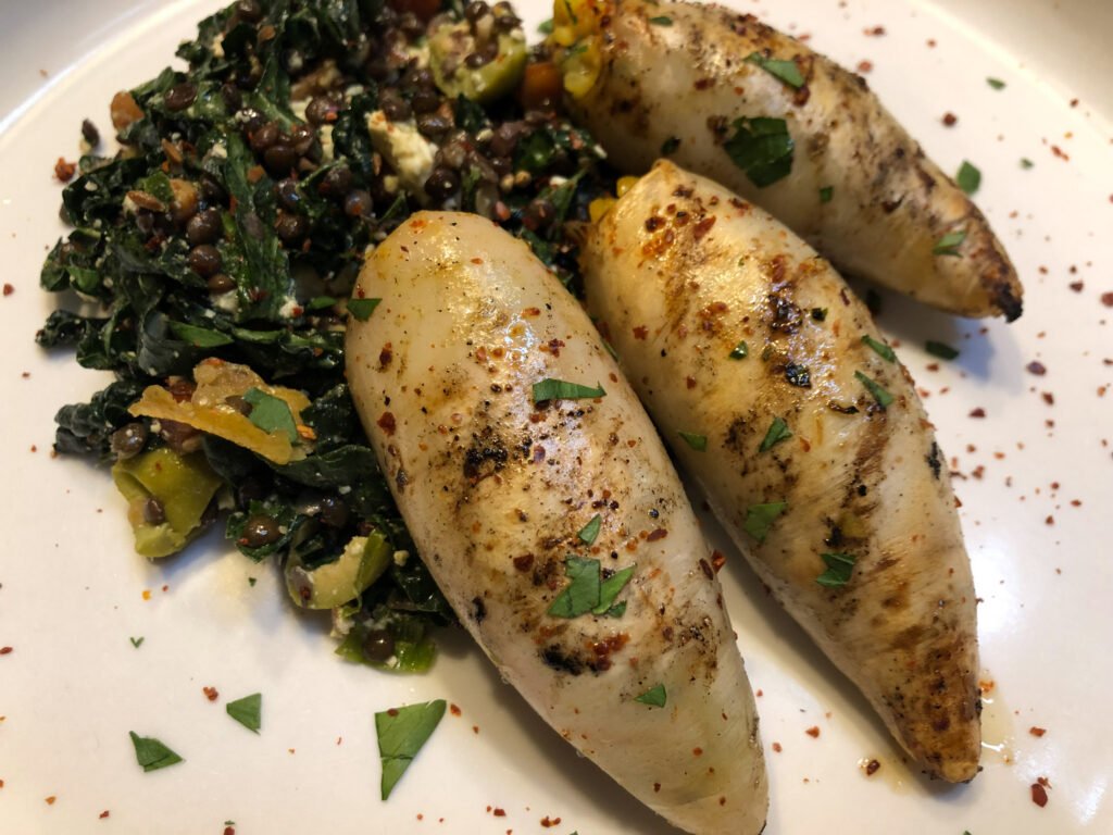 Three browned, herb-seasoned stuffed squid sit beside dark lentils and leafy greens on a white plate, creating a rustic, savory meal.