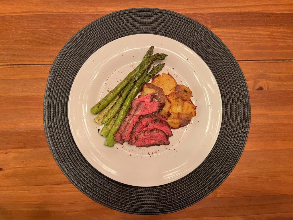 A white plate on a dark woven placemat holds sliced pink-centered roast beef, grilled asparagus, and golden roasted potatoes on a wooden table, presenting a warm meal.