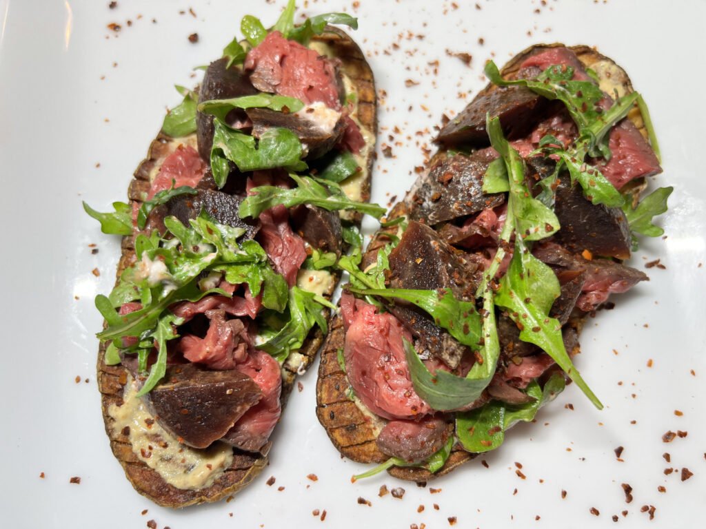 Overhead view of a gourmet steak salad on a white plate, with sliced medium-rare beef, grilled eggplant, fresh greens, and seasoning; inviting and rustic.