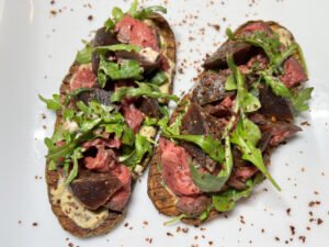 Overhead view of a gourmet steak salad on a white plate, with sliced medium-rare beef, grilled eggplant, fresh greens, and seasoning; inviting and rustic.