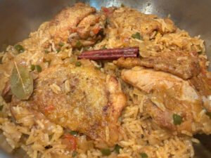 A pot of golden spiced rice topped with browned chicken pieces, a cinnamon stick, and bay leaf; the hearty, savory one-pot meal looks warm and inviting.