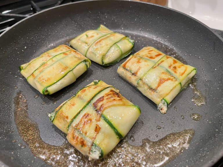 Four zucchini-wrapped parcels with golden-brown grill marks sizzle in oil in a dark skillet, suggesting a savory meal freshly cooked on the stovetop.